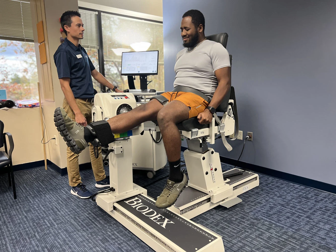 A man in an exercise chair undergoing leg-extension testing with a technician standing beside the machine in a clinic room.
