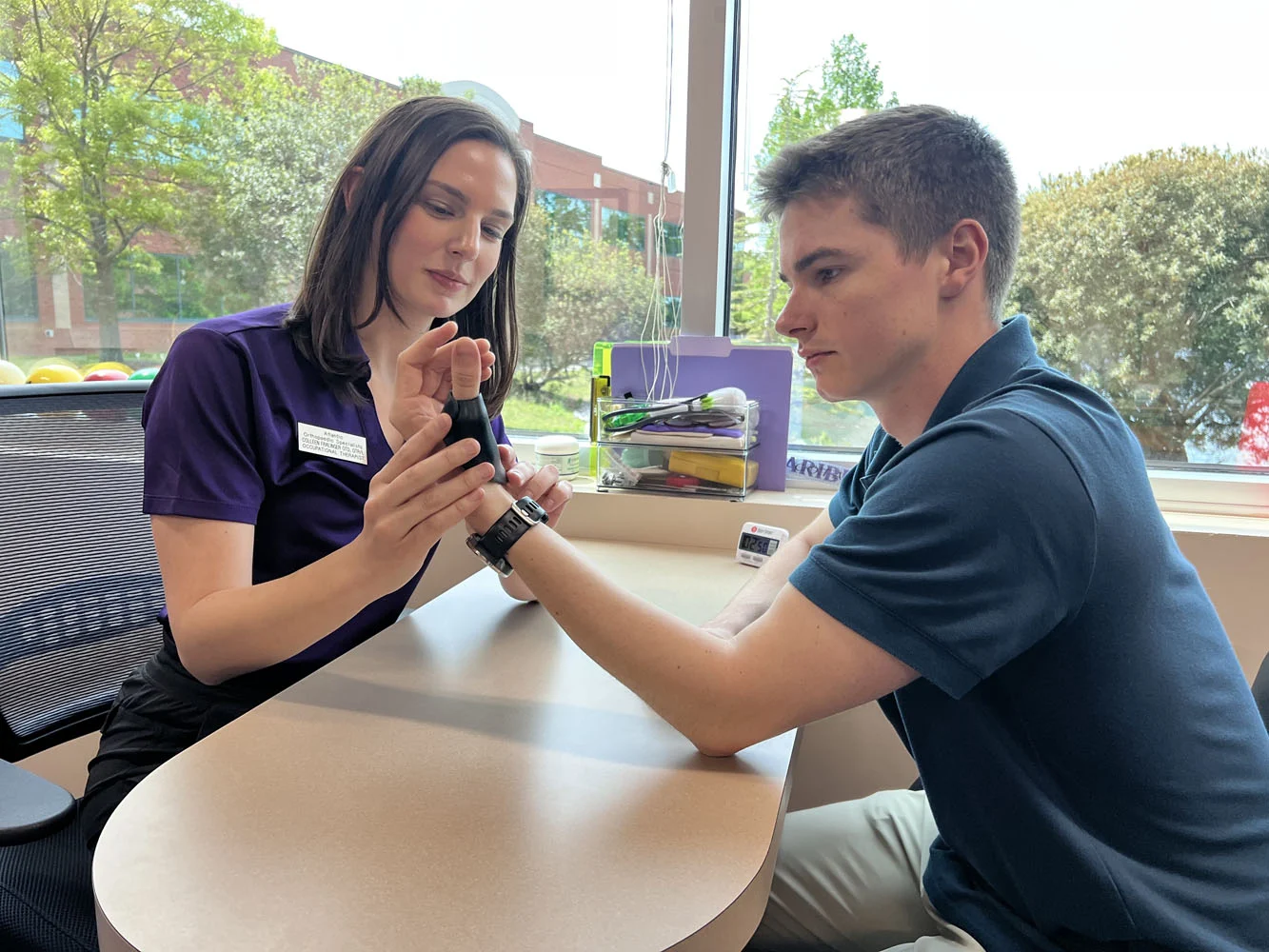 Female clinician in purple scrubs checks a male patient's wrist pulse at a clinic desk by a window.
