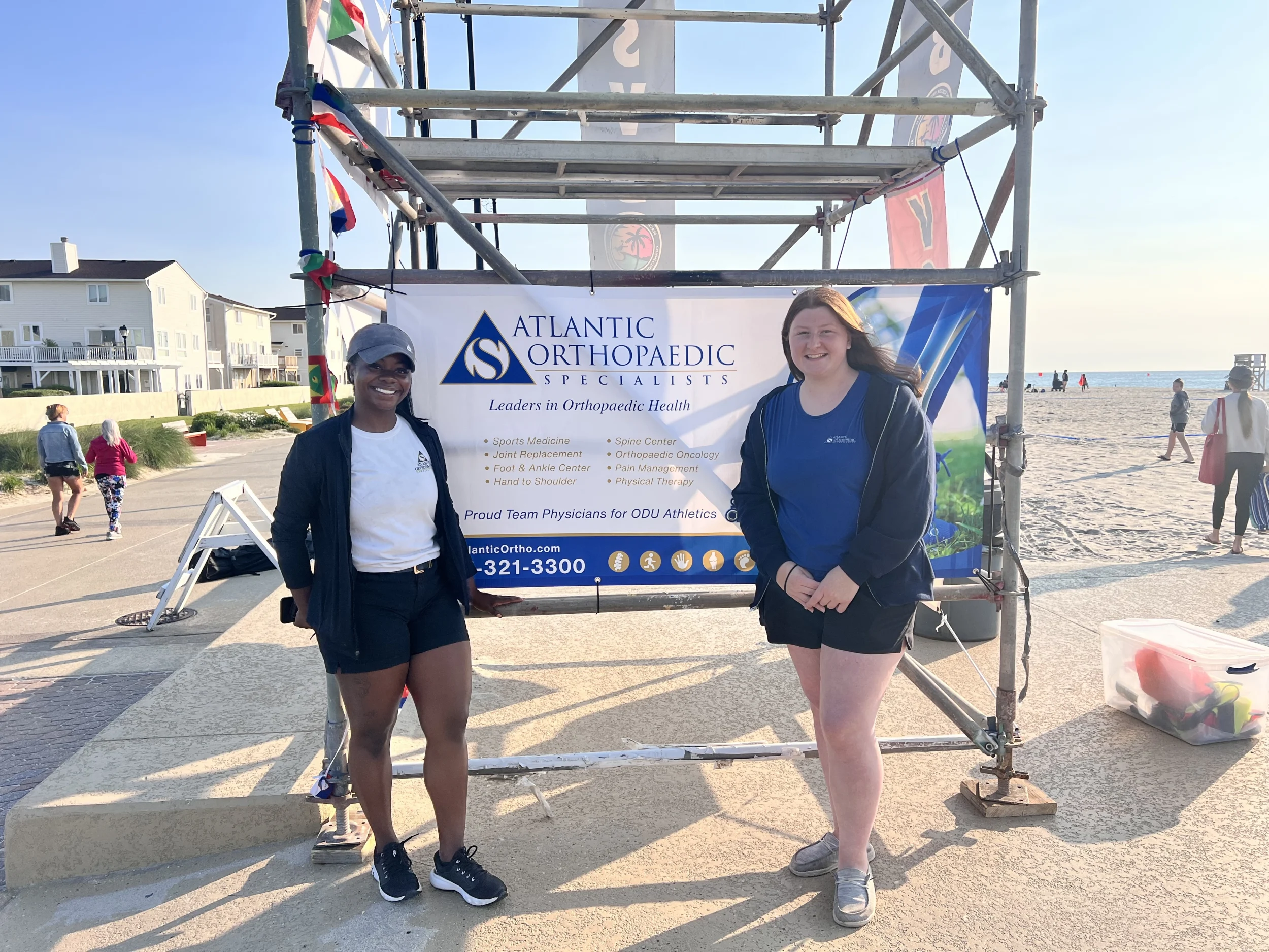 sand-soccer-2026 - Atlantic Orthopaedic Specialists - Atlantic Orthopaedic Specialists Two women pose in front of an Atlantic Orthopaedic banner at a beach event, with a metal scaffold and flags behind them on the boardwalk beach.