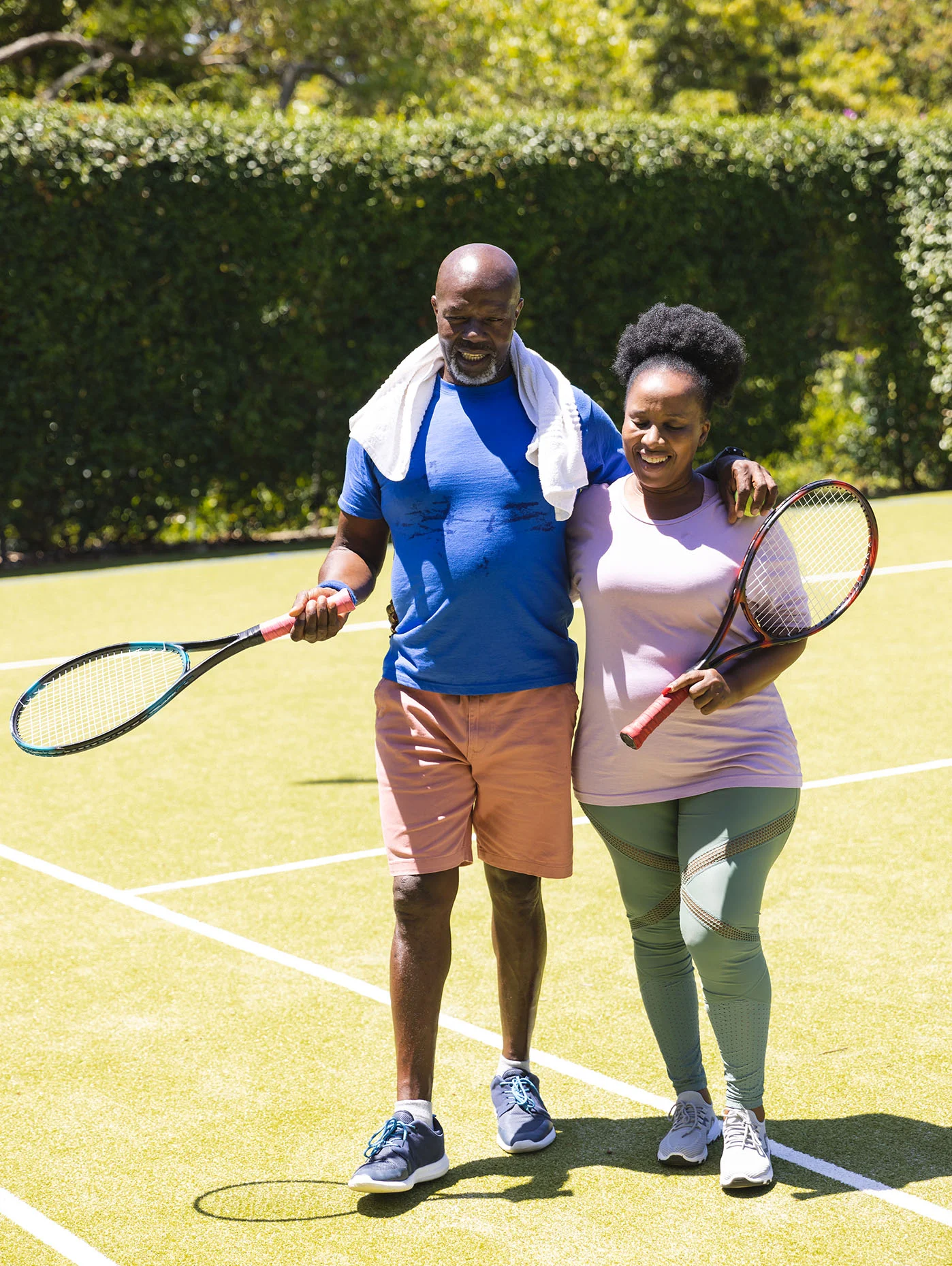 A smiling couple stands together on a tennis court with rackets, arms around each other after a game on a sunny day.