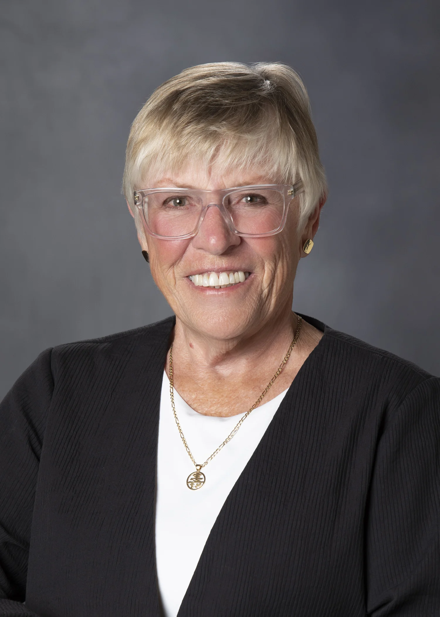 - Atlantic Orthopaedic Specialists Smiling older woman with short blonde hair and clear glasses, wearing a black blazer over a white top with a gold necklace, against a gray studio backdrop.